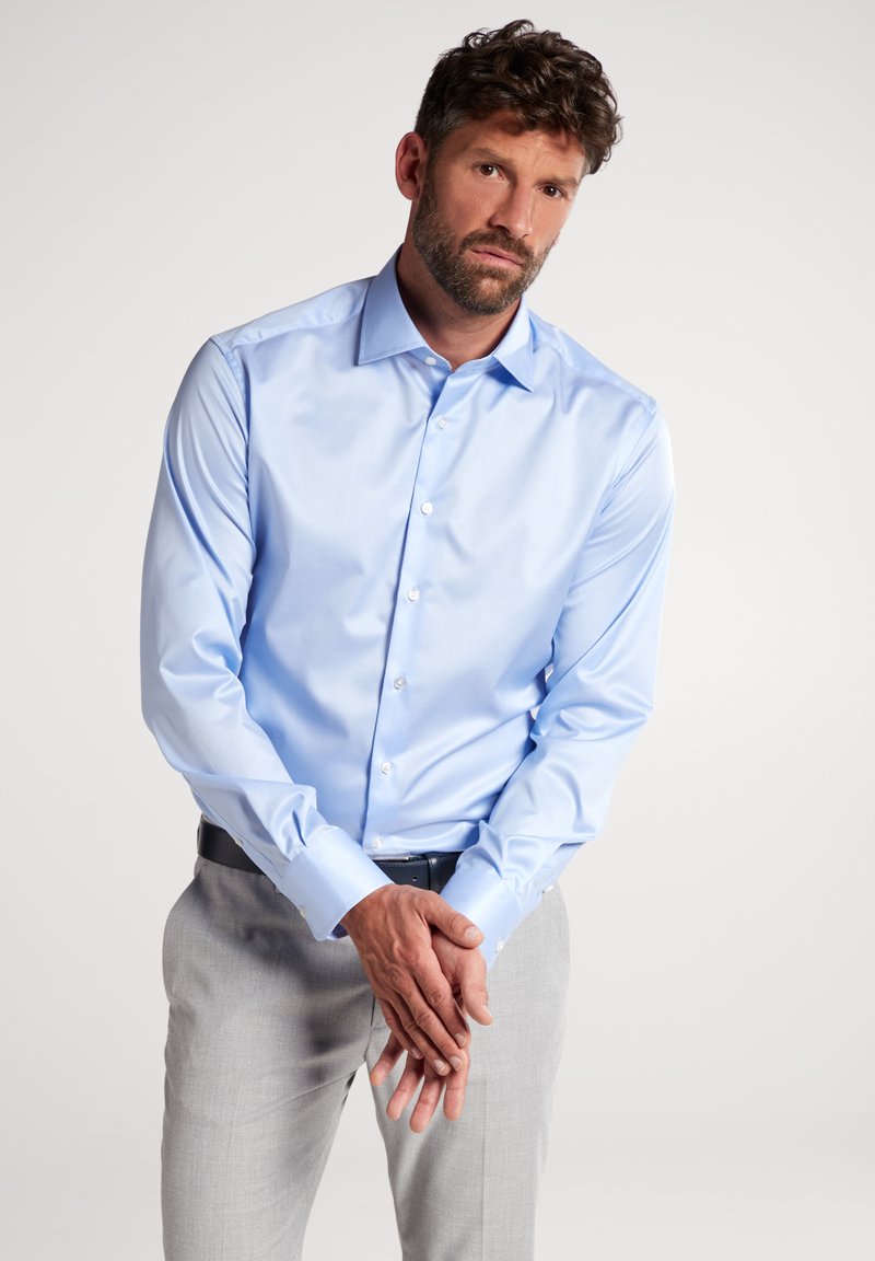 Camicia da uomo a maniche lunghe di colore azzurro chiaro in cotone morbido, con colletto classico e chiusura a bottoni, abbinata a pantaloni grigi chiari.