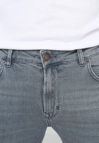 Jeans de mezclilla azul claro con cierre de botón, diseño de cinco bolsillos y un ligero desgastado. La tela tiene una textura suave con costuras reforzadas.