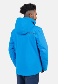 Chaqueta impermeable azul con capucha, con una textura suave y el logo negro "ROSSIGNOL" en la parte posterior. El diseño es simple y funcional.