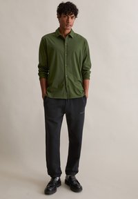 Roheline nööbitav särk kraega, kombineeritud mustade dressipükstega. Riietus on mugava lõikega ja sileda tekstuuriga.