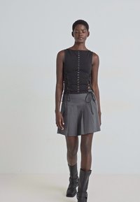 Top noir sans manches avec un design en dentelle sur le côté ; associé à une jupe plissée grise et des bottines noires. Tissus lisses avec une silhouette ajustée.
