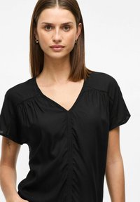 Blusa nera a maniche corte con scollo a V, vestibilità rilassata e sottili dettagli arricciati lungo il collo e le spalle. Tessuto dalla texture liscia.