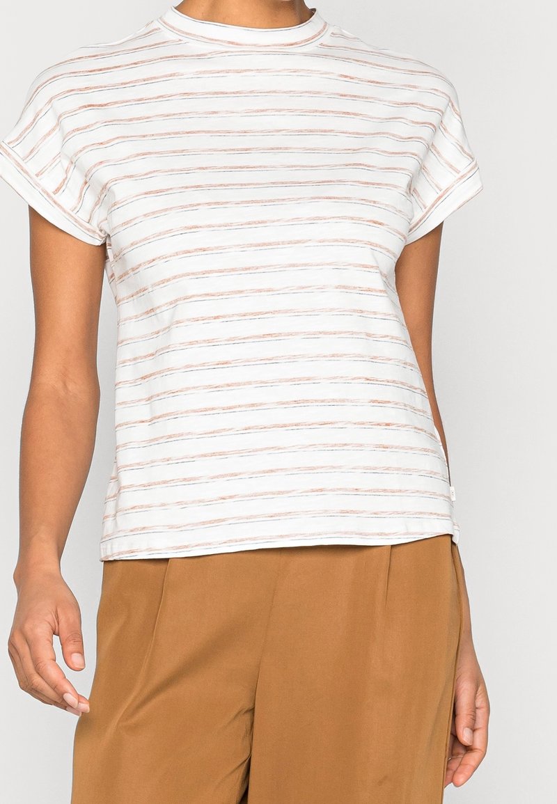 Femme portant un haut blanc à manches courtes avec de fines rayures horizontales marron et un pantalon marron ample sur un fond uni.