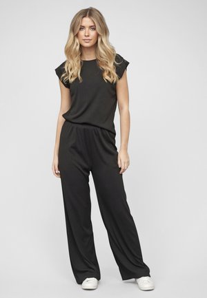 Blondynka stoi, ubrana w czarny bezrękawnik i luźne czarne spodnie oraz białe tenisówki na jednolitym tle.