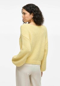 Rumena pletena jopica s širokimi rokavi in ribastim robom, ki ima teksturiran videz in sproščen kroj. Obuta je z svetlimi hlačami.