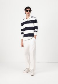 Marinblå och vit randig långärmad polo med krage, i kombination med vita byxor och ljusa sneakers; modell som bär runda svarta solglasögon.