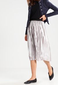 Metalliska plisserade culottbyxor i silver med en slät yta, ihop med en åtsittande svart topp och en marinblå jacka. Svarta spetsiga ballerinas fulländar looken.