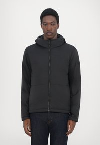 Schwarze Puffjacke mit Kapuze, mit einem Frontreißverschluss, horizontalen Nähten und elastischen Bündchen. Trägt sie über dunklen Jeans.