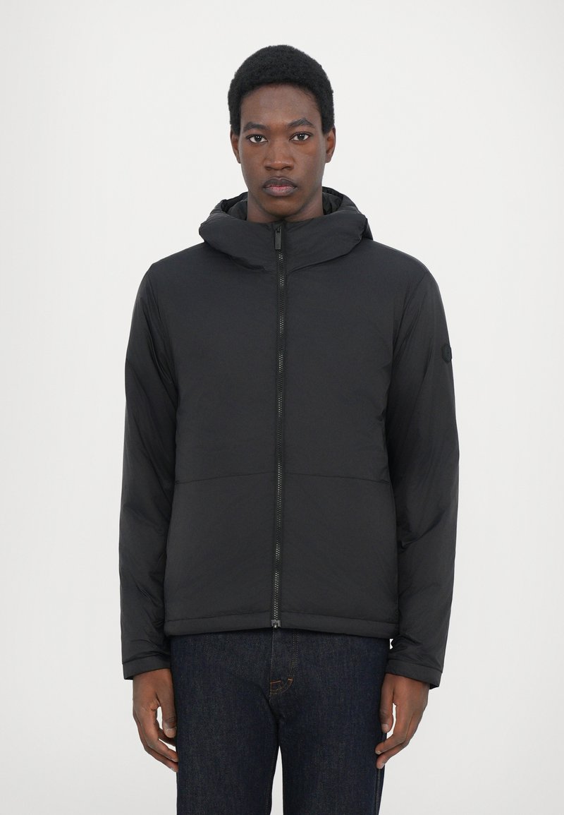 Schwarze Puffjacke mit Kapuze, mit einem Frontreißverschluss, horizontalen Nähten und elastischen Bündchen. Trägt sie über dunklen Jeans.