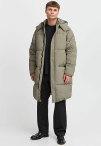 Polstret lang frakk i olivengrønn med hette, glidelås, sidelommer og kviltet design; kombinert med svarte bukser og sko.