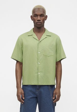 Muž s krátkymi blond vlasmi, oblečený v svetlozelenú košeľu s krátkym rukávom zapínanú na gombíky a modré džínsy, stojaci pred jednoliatym bielym pozadím.