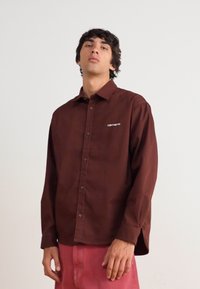 Καφέ πουκάμισο με κουμπιά από απαλή ύφανση και χαλαρή εφαρμογή, διαθέτοντας μικρό λευκό λογότυπο στο αριστερό στήθος. Μακριά μανίκια με κουμπιά στο μανίκι.