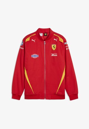 Czerwona kurtka wyścigowa Ferrari z żółtymi akcentami, logo Puma, zamkiem z przodu i wieloma naszywkami sponsorów na klatce piersiowej i rękawach.