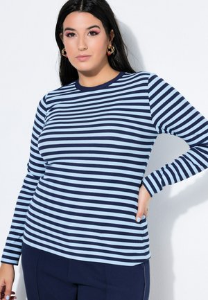 Kvinde med langt mørkt hår iført guld øreringe og en blå stribet langærmet skjorte, som poserer med den ene hånd på hoften.