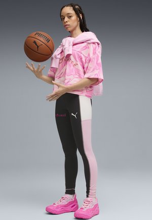 Giovane donna con capelli intrecciati che fa girare un pallone da basket Puma, indossando abbigliamento sportivo rosa e nero e scarpe da ginnastica rosa acceso su uno sfondo grigio.