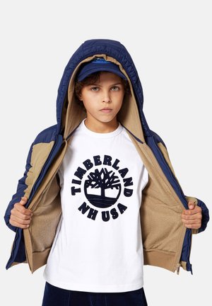 Blouson matelassé marine avec doublure beige, ouvert pour révéler un t-shirt blanc avec un logo d'arbre circulaire noir. Casquette bleue avec logo.