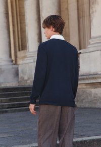 Tumesinine pika varrukaga kootud top valge kraega, kombineeritud pruunide ruudulise mustriga pükstega, vaadates selja tagant kivisammaste taustal.