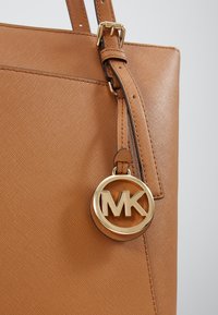 Sac à main en cuir texturé tan avec une boucle dorée et un charme rond au logo MK suspendu de la bandoulière sur un fond gris.