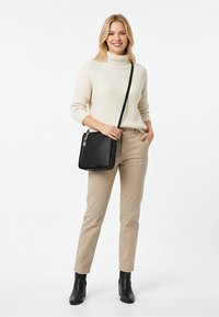 Donna che indossa un maglione dolcevita color crema, pantaloni beige, stivaletti neri alla caviglia e porta una borsa a tracolla nera, sorridente, con una mano in tasca.