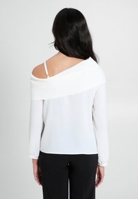 Blusa bianca a maniche lunghe con design off-shoulder, caratterizzata da una texture liscia e un leggero brillante. Il retro presenta un dettaglio di cucitura e una leggera caduta.