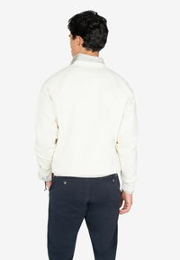 Sudadera blanca con un cuello redondo, tela texturizada y puños acanalados, combinada con pantalones oscuros que tienen bolsillos traseros y detalles de metal.