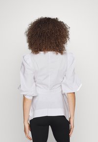 Blusa bianca con maniche a palloncino, vita aderente e tessuto texturizzato. Presenta un retro dritto e una leggera lucentezza, abbinata a leggings neri.