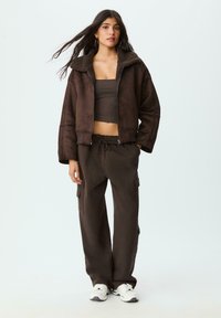 Giubbotto bomber in finta pelle scamosciata marrone con colletto in shearling, abbinato a pantaloni cargo marroni con tasche laterali e sneakers bianche.