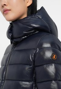 Veste rembourrée marine avec une finition brillante, design matelassé, col montant et patch de logo circulaire sur la manche gauche. Fermeture zippée à l'avant.
