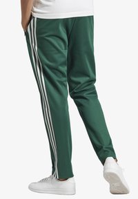 Gröna sweatshirtbyxor i slät tyg med tre vita ränder längs varje ben. Kombinerade med vita sneakers, visad bakifrån.