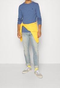 Blå långärmad skjorta, gul tröja knuten i midjan, ljusblå jeans, grå sneakers med vita ränder och gula strumpor.