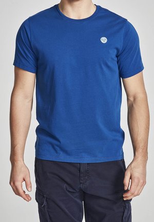 Mann trägt ein königsblaues kurzärmeliges T-Shirt mit einem kleinen weißen runden Logo auf der linken Brust, kombiniert mit dunkelblauen Cargo-Shorts.