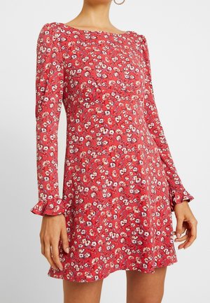 Frau trägt ein rotes, langärmeliges Blumenkleid mit weißen und pinken Blumen, mit gerafften Bündchen und tailliertem Schnitt.