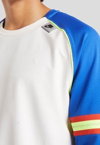 Biała koszula z krótkim rękawem z niebieskimi teksturowanymi rękawami, neonowo zielonym wykończeniem i czerwonymi akcentami. Logo Nike umieszczone na lewym ramieniu.