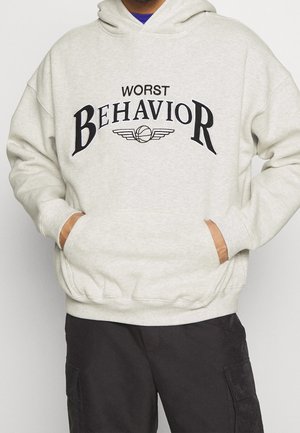 Personne portant un sweat à capuche gris clair avec le texte "WORST BEHAVIOR" et un graphisme de basket ailé sur la poitrine, mains dans la poche avant.