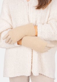 Person, der beigen Strickhandschuhe über einem cremefarbenen, strukturierten Pullover trägt, mit verschränkten Armen auf der Taille.
