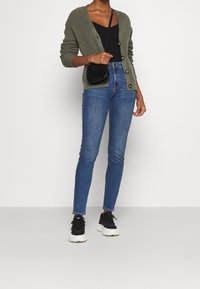 Cárdigan de punto verde oliva con botones, camiseta negra, jeans de tiro alto azules y zapatillas negras con suela blanca gruesa.