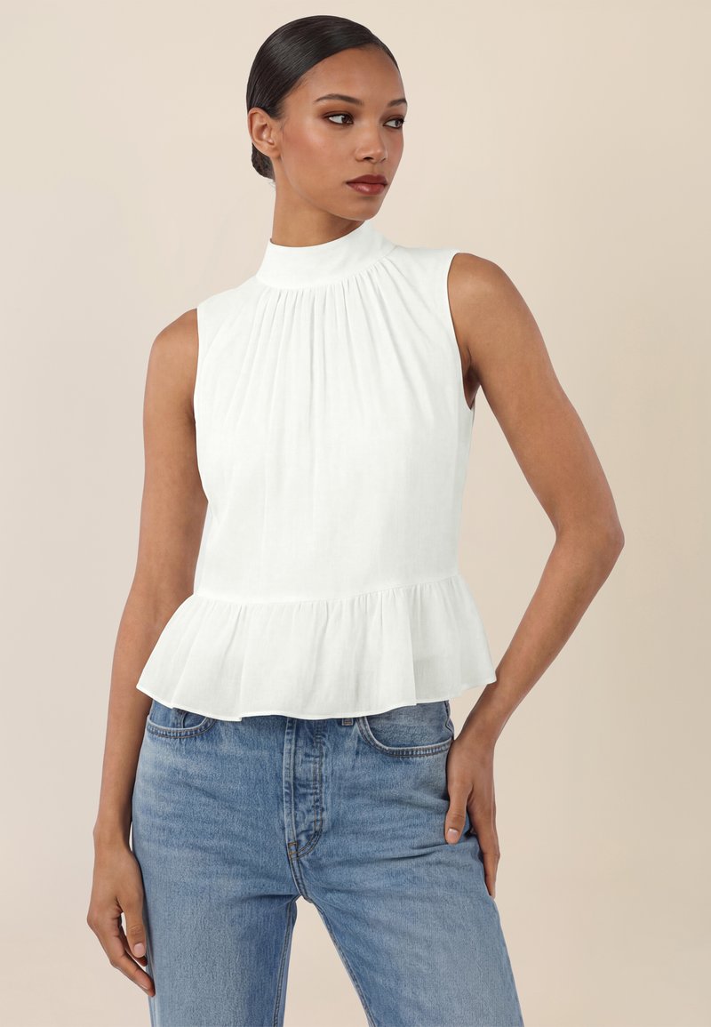 Mujer con el cabello recogido en un moño, vestida con una blusa blanca sin mangas con volante en el dobladillo y cuello alto, combinada con jeans azul claro, de pie contra un fondo beige.