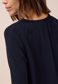Camicia a maniche lunghe blu navy con tessuto morbido e liscio. Dettaglio del collo arricciato e vestibilità rilassata, che mette in risalto un design semplice con una sottile eleganza.
