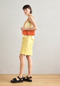 Geeltje mouwloze top en een geel rok tot de knie, gecombineerd met zwarte sandalen en een oranje handtas met een sluitingdetail.