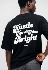 Jeune homme portant un t-shirt noir avec le texte blanc "Hustle Hard, Shine Bright" dans le dos, tenant le bord du t-shirt d'une main.