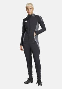 Veste de sport noire avec accents gris, fermeture éclair devant et poches latérales, associée à des leggings noirs avec des rayures réfléchissantes et une coupe ajustée.