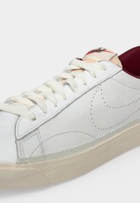 Λευκό δερμάτινο sneaker με λευκά κορδόνια, διάτρητο σχέδιο Swoosh, μπεζ ελαστική σόλα και μπορντό εσωτερική επένδυση ορατή κοντά στην περιοχή του αστραγάλου.