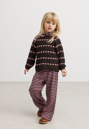 Jeune fille aux cheveux blonds portant un pull noir avec des cœurs roses et un pantalon rayé rose et noir, debout sur un fond uni.