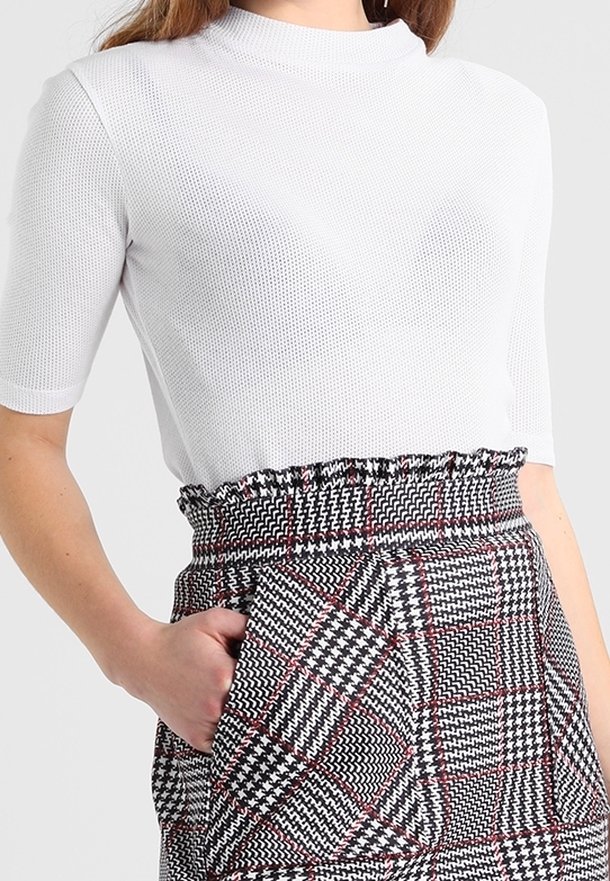 Femme portant un haut en maille blanc à manches courtes, rentré dans une jupe à carreaux noir, blanc et rouge taille haute, avec une ceinture volantée et une main dans la poche.