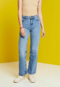 Jeans de mezclilla azul claro con un corte de cintura alta, patas rectas y dobladillos deshilachados. Combinados con zapatillas blancas sobre un fondo amarillo.