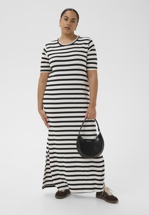 Vrouw draagt zwart-wit gestreepte maxi-jurk met korte mouwen, houdt een zwarte halvemaanvormige handtas vast en draagt donkere sneakers met witte veters.