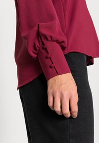 Burgundská blůza s nafouknutými rukávy, knoflíkovými manžetami s třemi dekorativními textilními knoflíky a zvlněným lemem, kombinovaná s tmavými džíny.