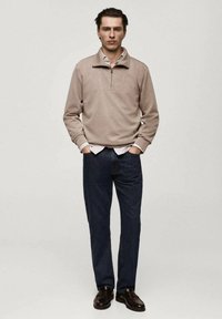 Pullover beige con zip al collo, in misto cotone, maniche lunghe, abbinato a jeans a gamba dritta blu scuro e scarpe in pelle marrone.