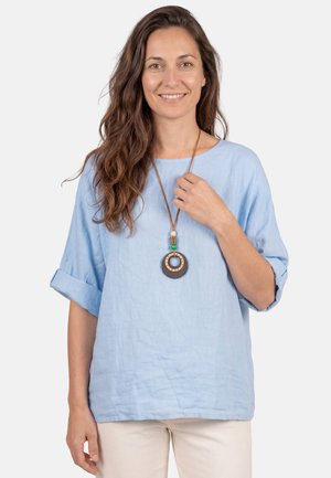 Frau mit langem, welligem Haar, die ein hellblaues Top mit hochgekrempelten Ärmeln und beige Hosen trägt und eine runde Halskette mit Holzperlen hält.