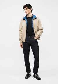 Νέος άνδρας με σγουρά μαλλιά, φορώντας μπεζ μπουφάν με μπλε επένδυση, μαύρο πουκάμισο, σκούρα τζιν και μαύρα μοκασίνια, στέκεται με το ένα χέρι στην τσέπη.